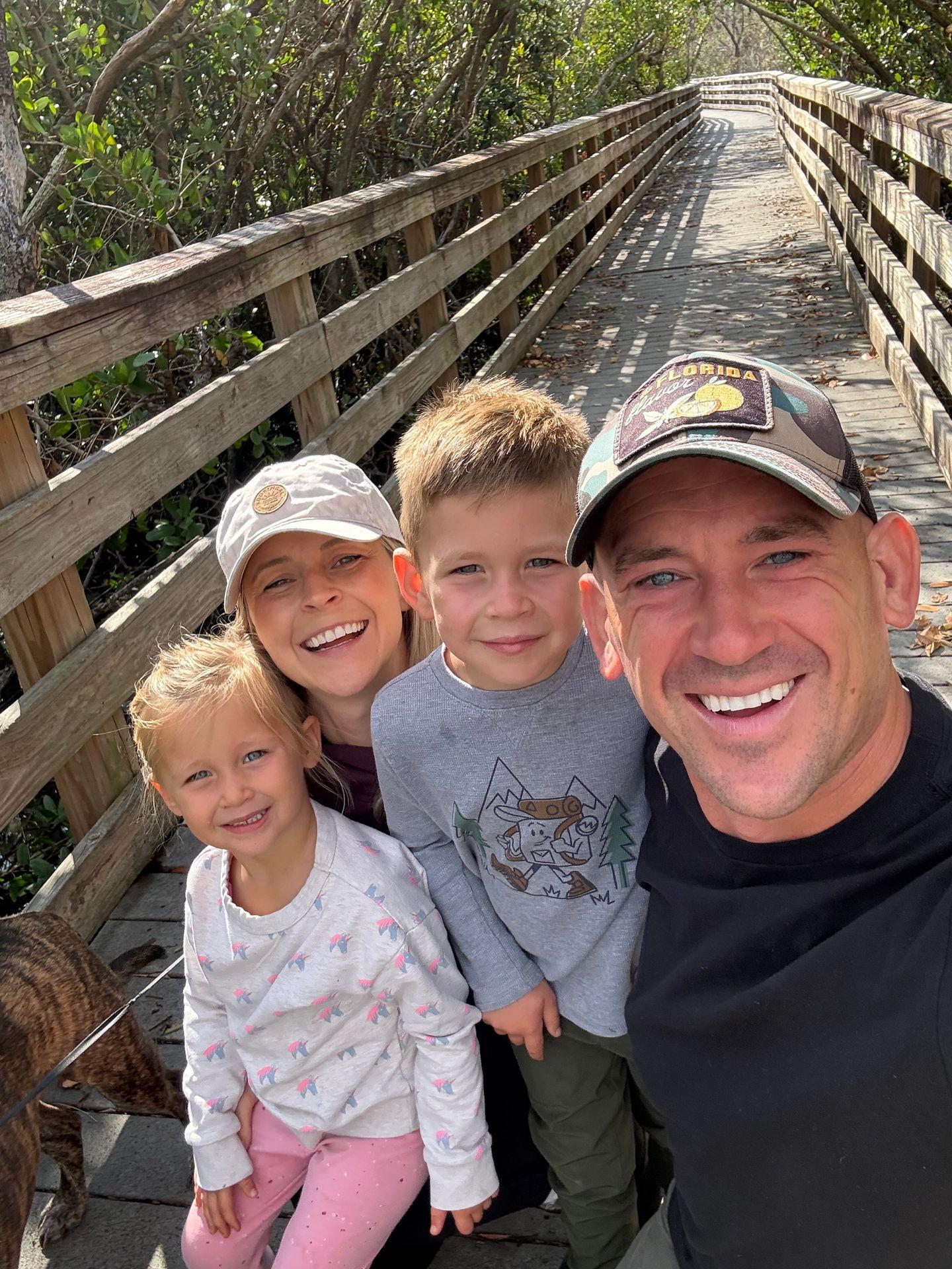 Smiling family of four taking a selfie on a wooden walkway surrounded by greenery.